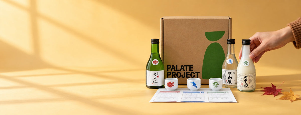 A hand reaches for one of three sake mini-bottles beside the Sake Flight gift box, three cups and a tasting mat on a warm yellow backdrop in window light, with two autumn maple leaves